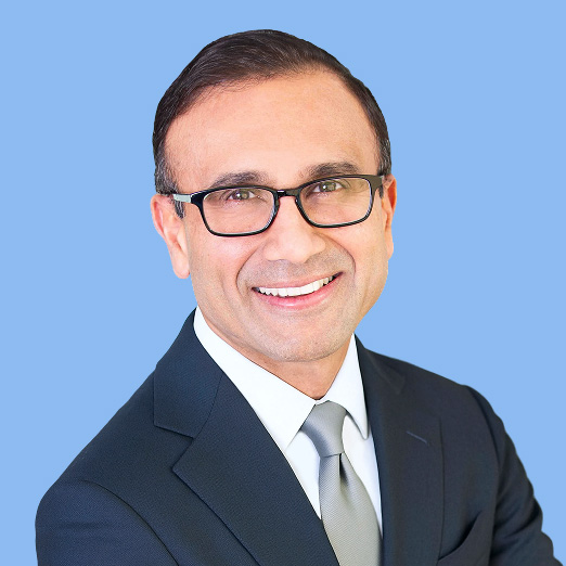 Professional headshot smiling man in a dark suit and glasses against a light blue background