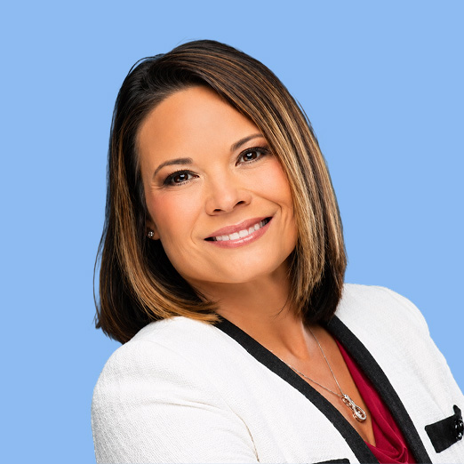 Smiling woman with shoulder length highlighted brown hair white blazer burgundy top against a blue background