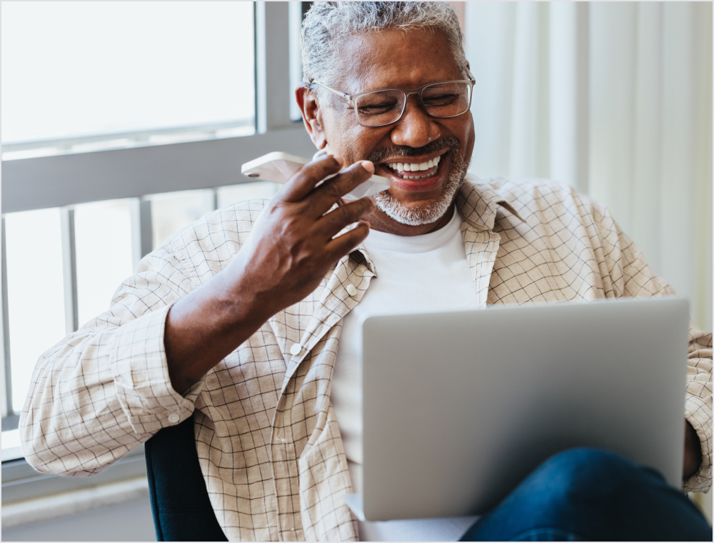 Smiling older man with gray hair and glasses holds a tissue to his mouth while using a laptop