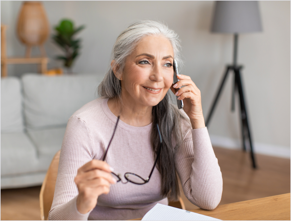 Older woman with gray hair talking on a mobile phone smiling holding her glasses in one hand at a home desk